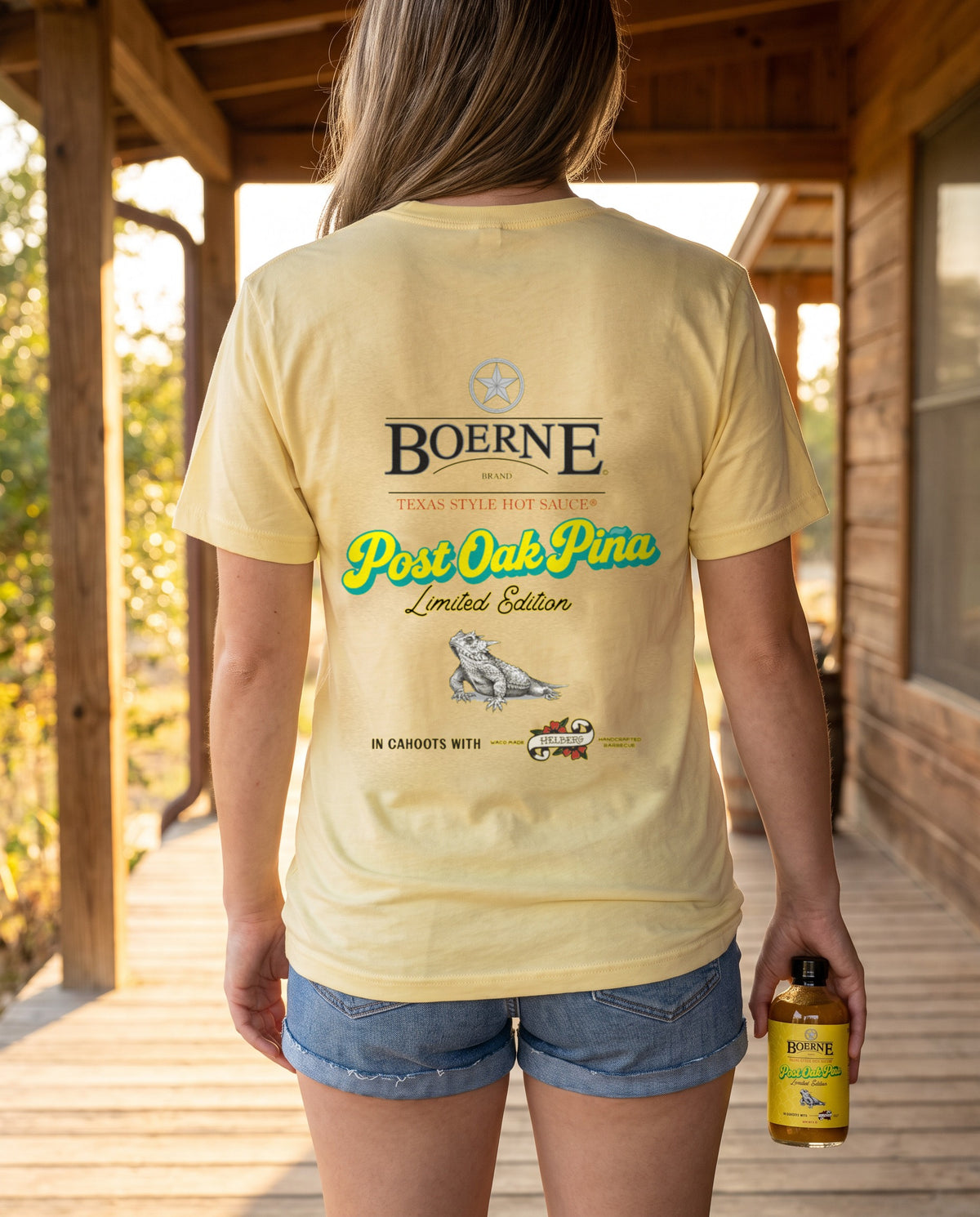 Post Oak Pina Tee — Pale Yellow Triblend, model wearing back print on a Texas porch, holding Post Oak Pina bottle