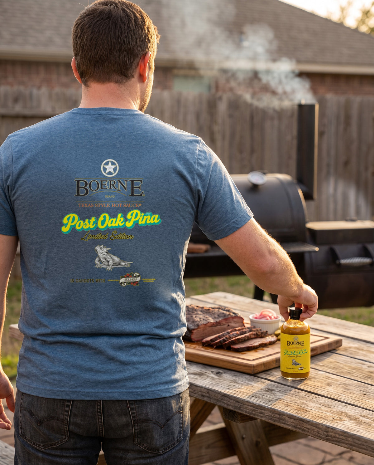 Post Oak Pina Tee — Denim Triblend, model reaching for Post Oak Pina bottle on backyard smoker table with sliced brisket